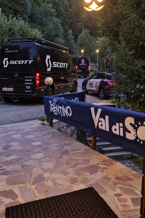 Une zone pavée, délimitée par une barrière bleue sur laquelle est inscrit le texte « Val di Sol », entourée de véhicules stationnés et d’arbres.