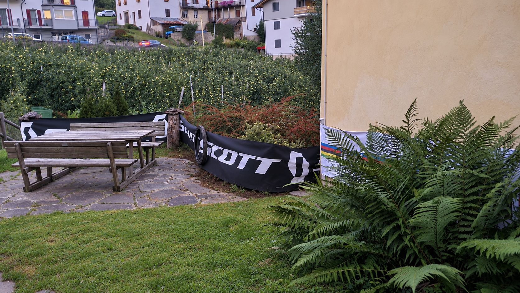 Un jardín con un banco de madera y un estandarte negro con letras blancas.
