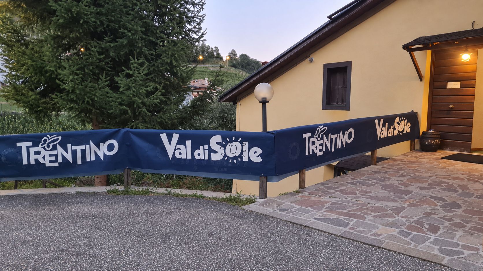 Une entrée d’immeuble avec un chemin de pierre menant à une porte, une banderole bleue portant les inscriptions « Val di Sole » et « Trentin », et un lampadaire.
