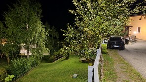 Une vue nocturne d’un quartier résidentiel, avec une voiture garée, une pelouse bien entretenue et une maison dont la véranda est visible.