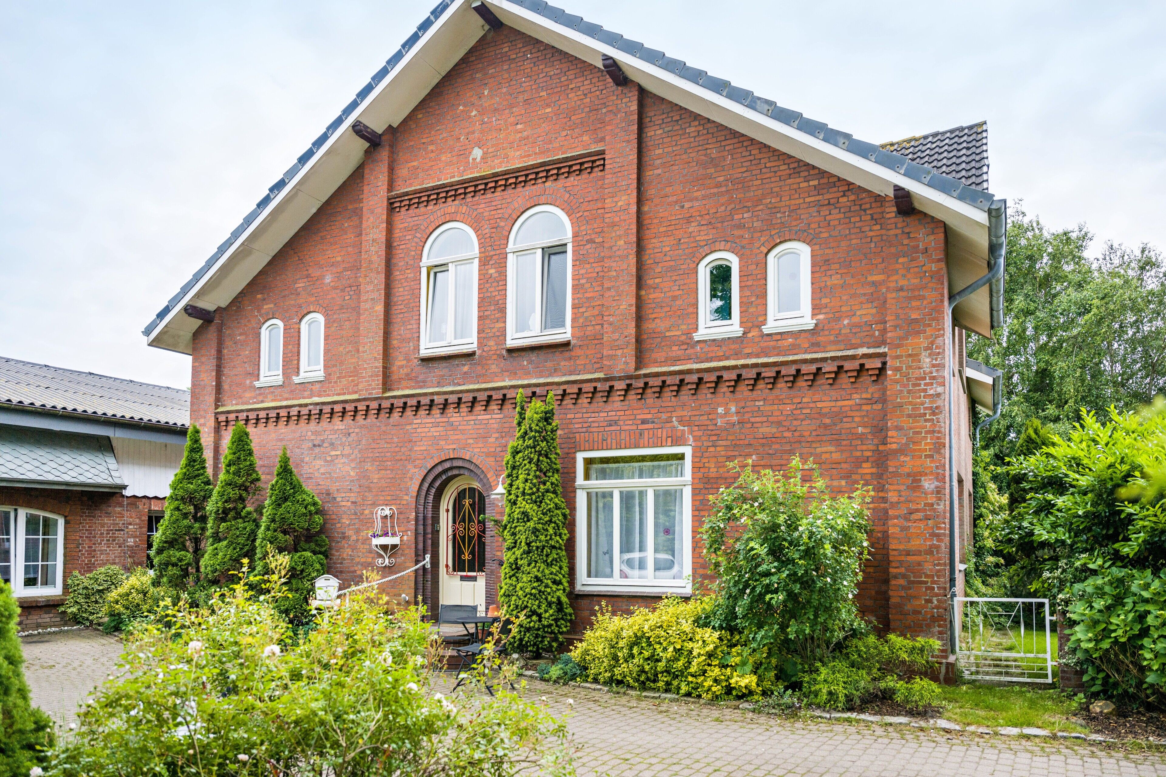 A red brick building with a pitched roof, multiple windows, and a small garden area with greenery.