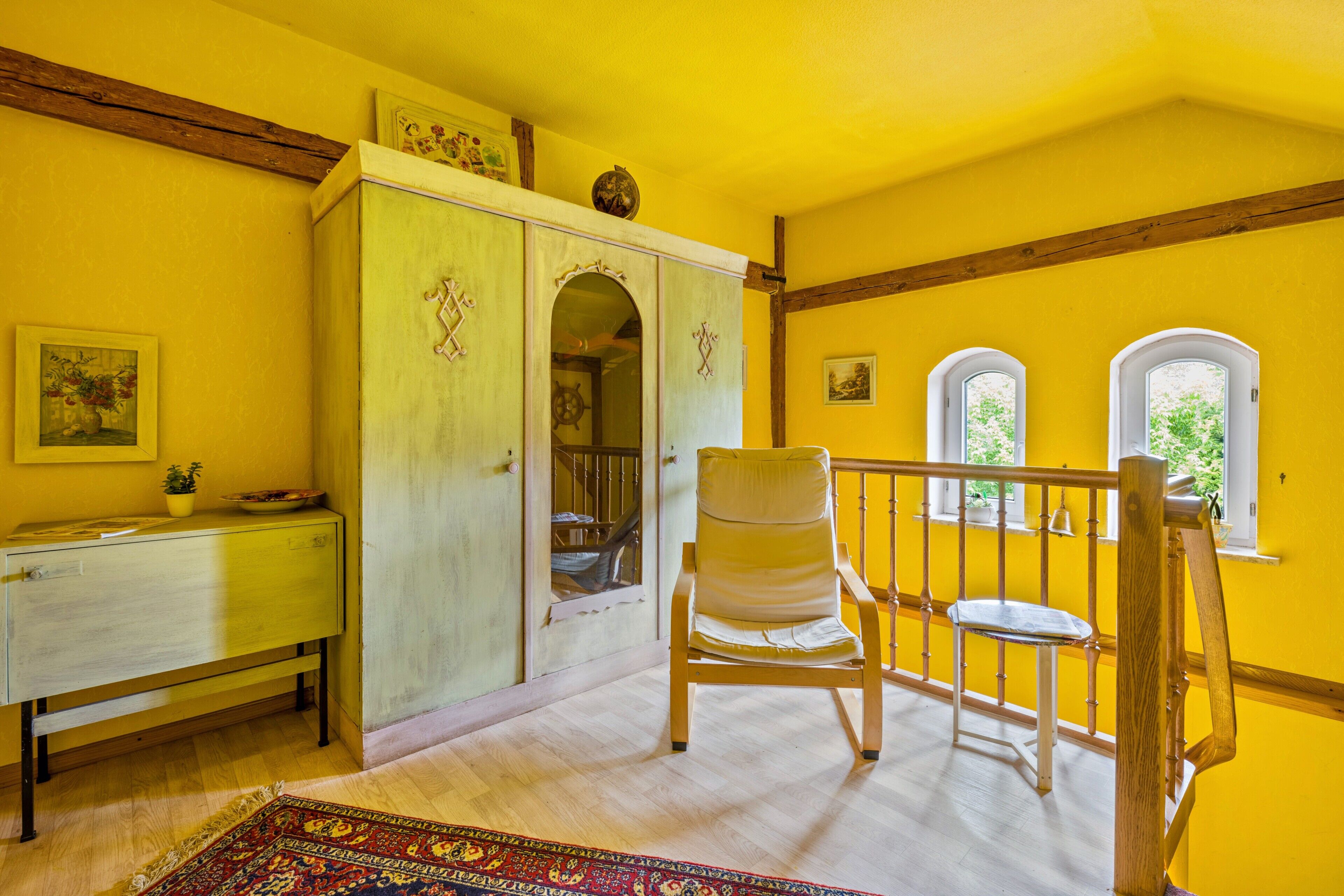 Une pièce avec une chaise en bois, un bureau, une armoire et un escalier, le tout dans un décor comprenant un mur jaune et des poutres en bois.