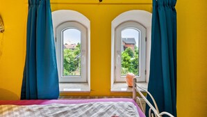 A room with yellow walls, a bed with a checkered bedspread, two windows with arched tops, and blue curtains. - Room 'Single Room 2' with Wi-Fi (Neuenkirchen)