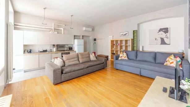 A modern living room with a grey sofa, a blue sofa, and a wooden coffee table. There is a kitchen area with white cabinets and a refrigerator. - San Gregorio House (Milano)