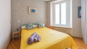 A bedroom with a yellow bedspread, a wooden nightstand, a framed picture on the wall, and a window with curtains. - San Gregorio House (Milano)