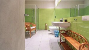 A bathroom with a green tiled wall, a white sink, a white bathtub, a wicker chair, and a wooden bench. - San Gregorio House (Milano)