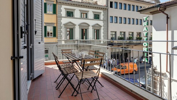 Un balcón con una mesa y sillas, con vista a una calle con edificios y un automóvil.
