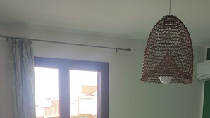 Une pièce avec un lit vert, une table et une chaise, une lampe suspendue et une vue sur des immeubles par la fenêtre.