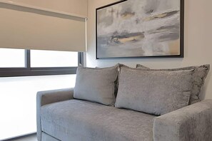 Ein modernes Wohnzimmer mit einem grauen Sofa, einem großen abstrakten Gemälde und einem Fenster mit Jalousien.
