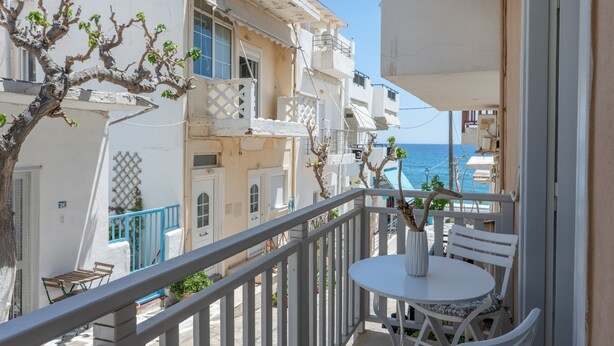 A balcony with a white table and chairs, overlooking a coastal area with buildings and the sea.