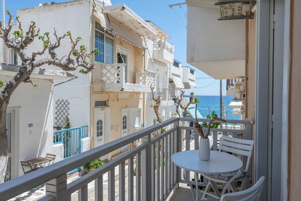 A balcony with a white table and chairs, overlooking a coastal area with buildings and the sea.
