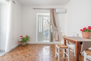 A room with a tiled floor, a white chair, a wooden table with a cloth, a potted plant, and a door leading to an outdoor area.