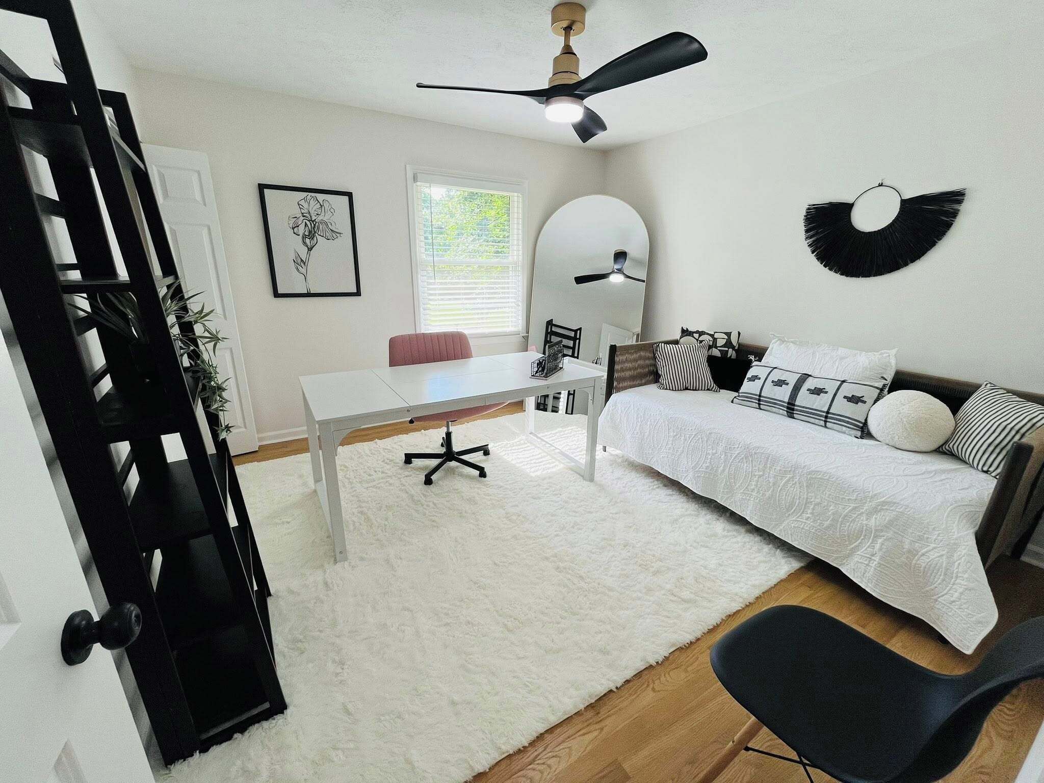 Camera da letto moderna con un letto bianco, una scrivania e una sedia. C'è una ventola a soffitto, una finestra con persiane e uno specchio a parete.