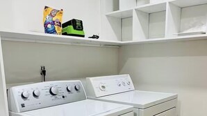 Un cuarto de lavandería con lavadora y secadora de carga frontal, un estante con una bolsa de detergente y un enchufe.