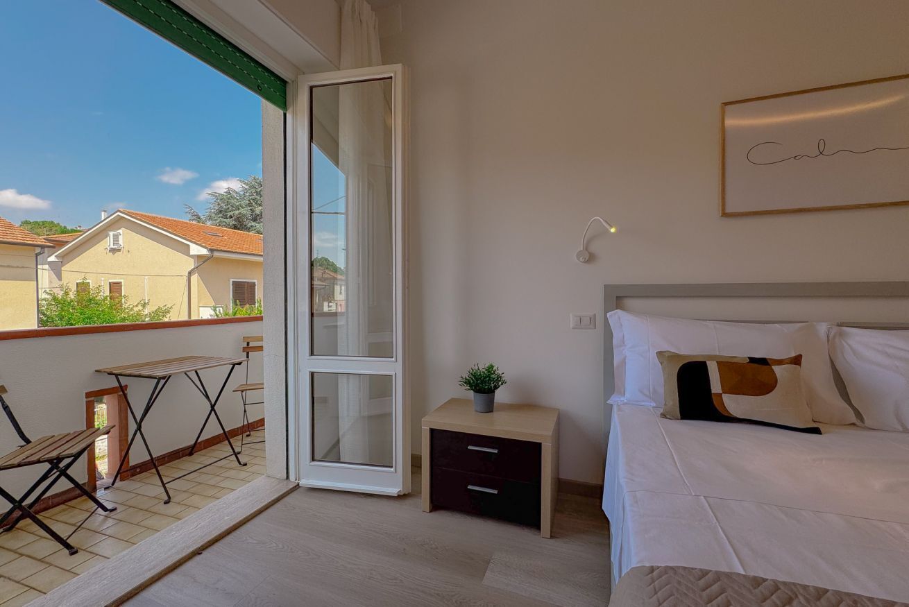 Una camera da letto con un letto, un comodino, un tavolino, una sedia, una pianta in vaso e una porta scorrevole in vetro che dà su un balcone esterno.