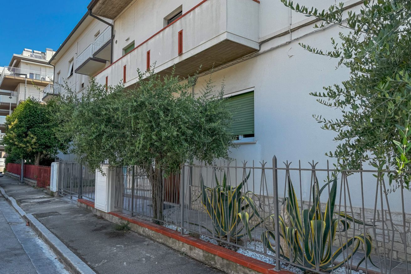 Um edifício residencial com varandas, portão fechado e uma fileira de árvores.