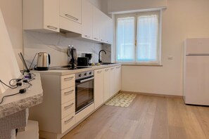 Ett kompakt kök med vita skåp, en inbyggd ugn, en kaffebryggare och ett fönster med tunna, genomskinliga gardiner.