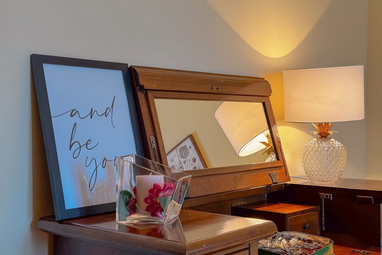 A room with a wooden dresser, a mirror, a framed sign, a lamp, and a vase.