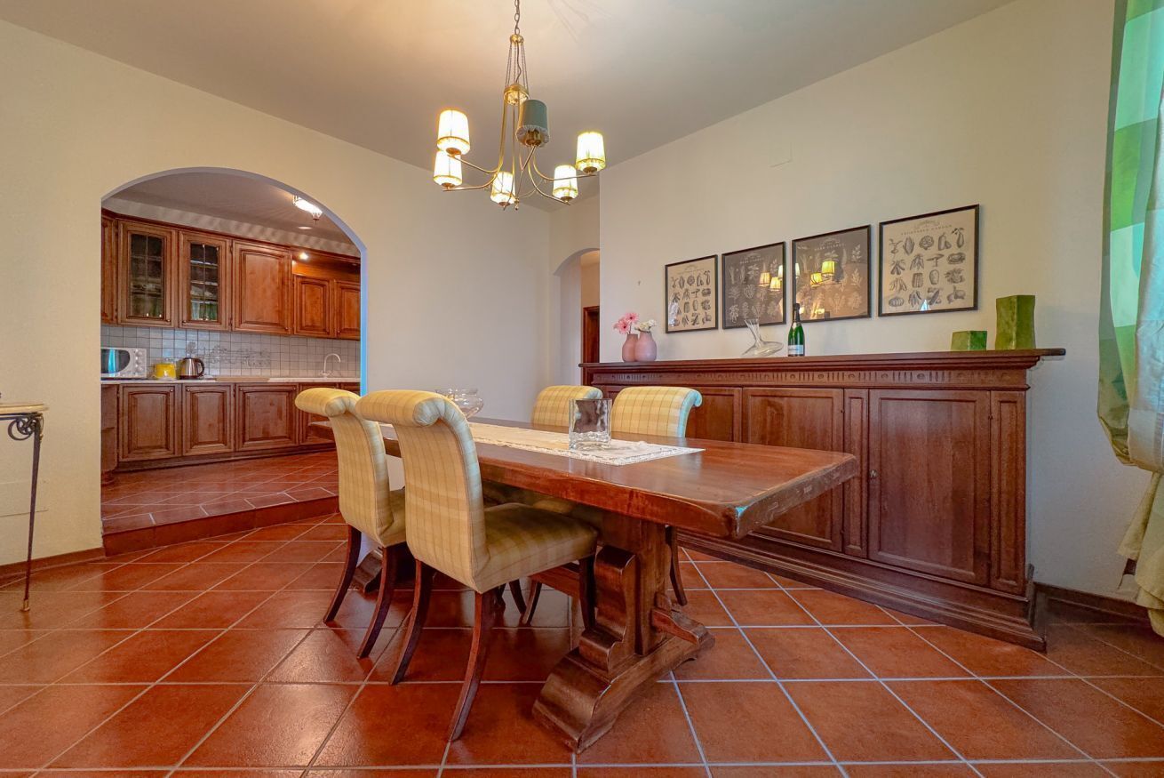 A dining area with a wooden table and chairs, a kitchen in the background, and a chandelier.