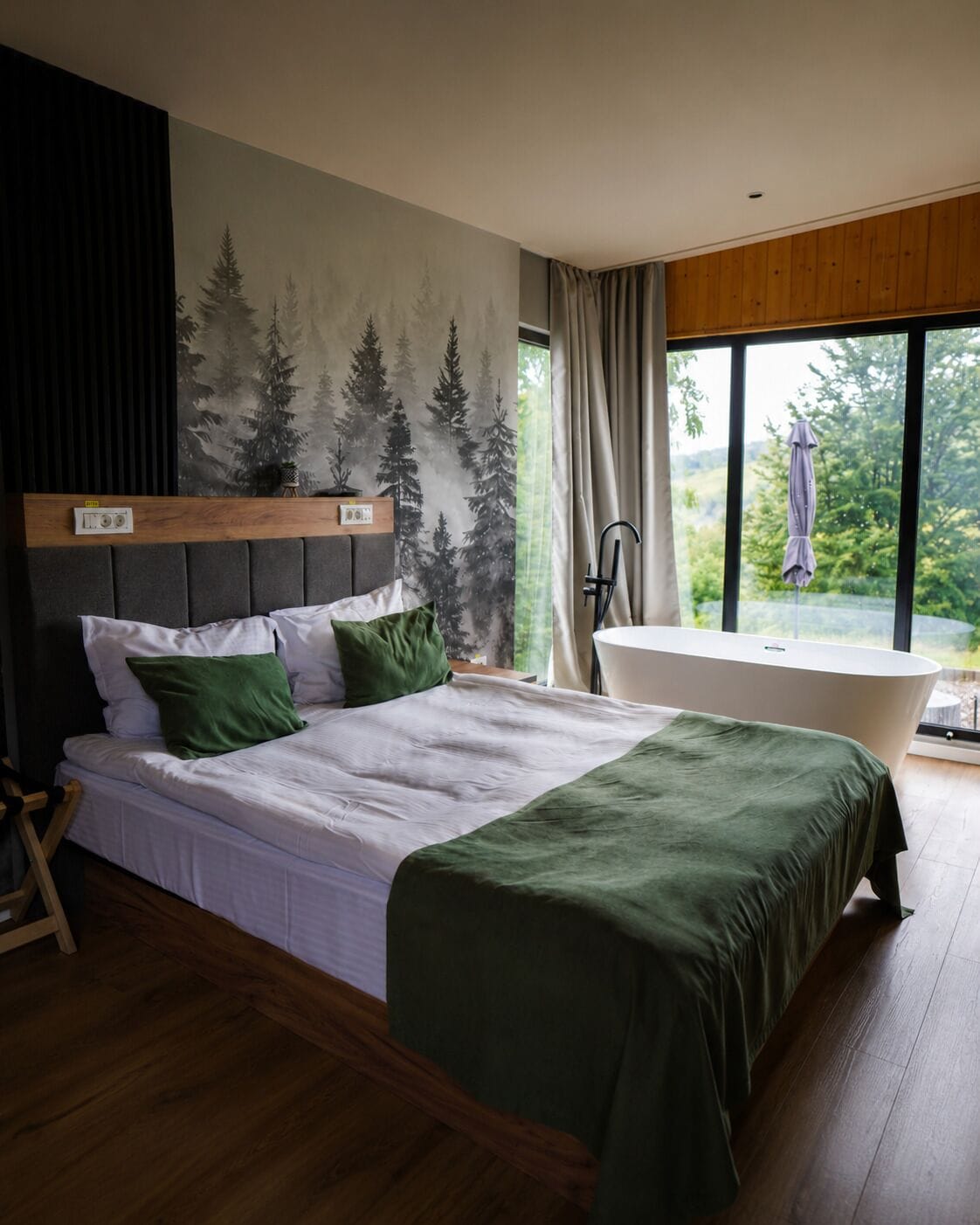 Un dormitorio con cama, un ventanal grande, una bañera y un mural temático de bosque.