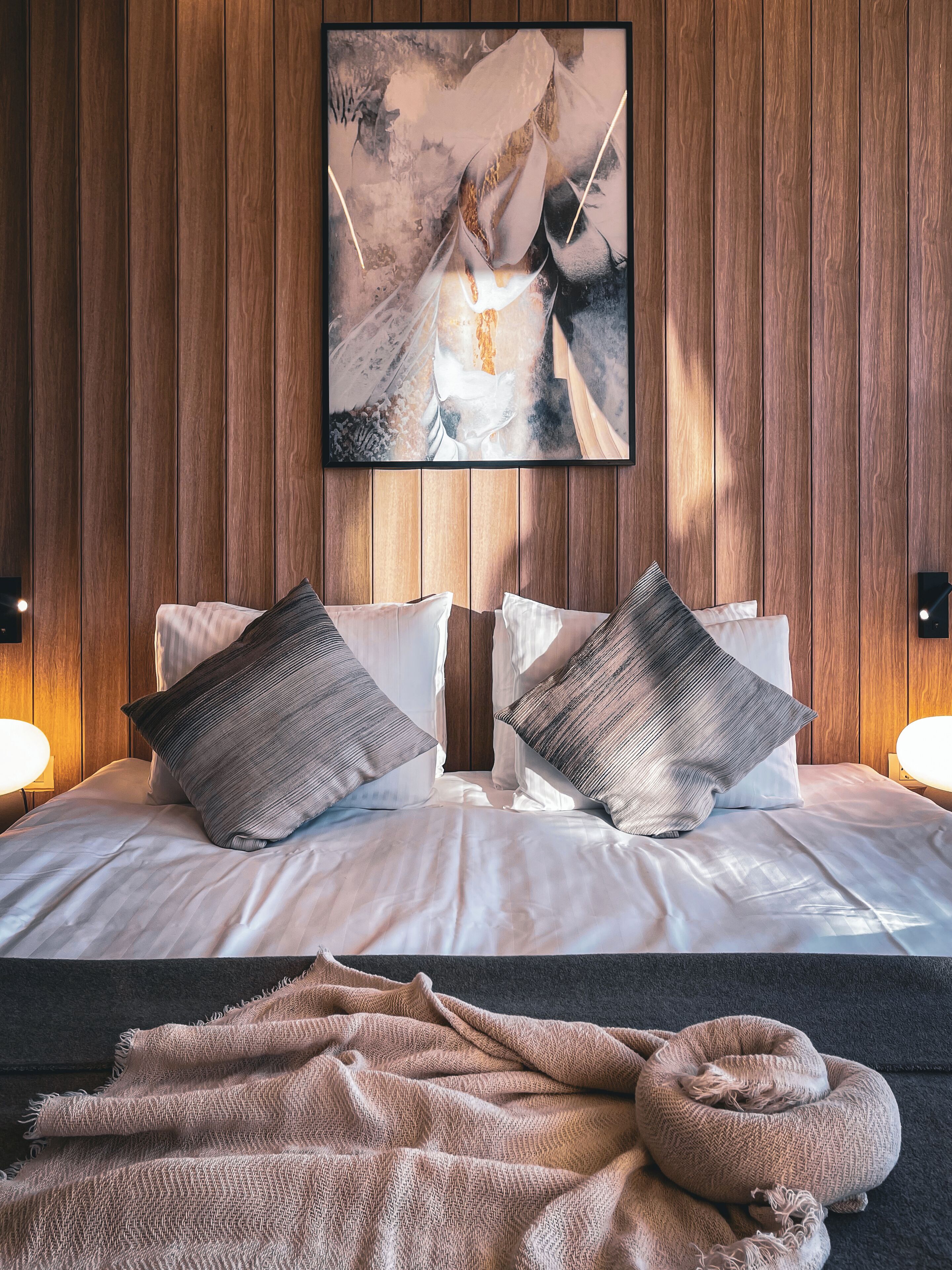 Una cama bien hecha con dos almohadas, una colcha y un cabecero frente a una pared de madera.