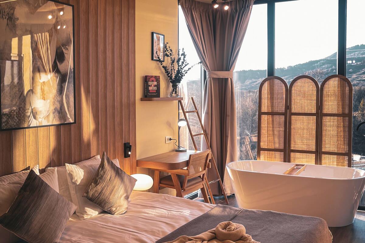 Een slaapkamer met een groot bed, een bureau, een stoel, een lamp, een boekenkast en uitzicht op het groen.