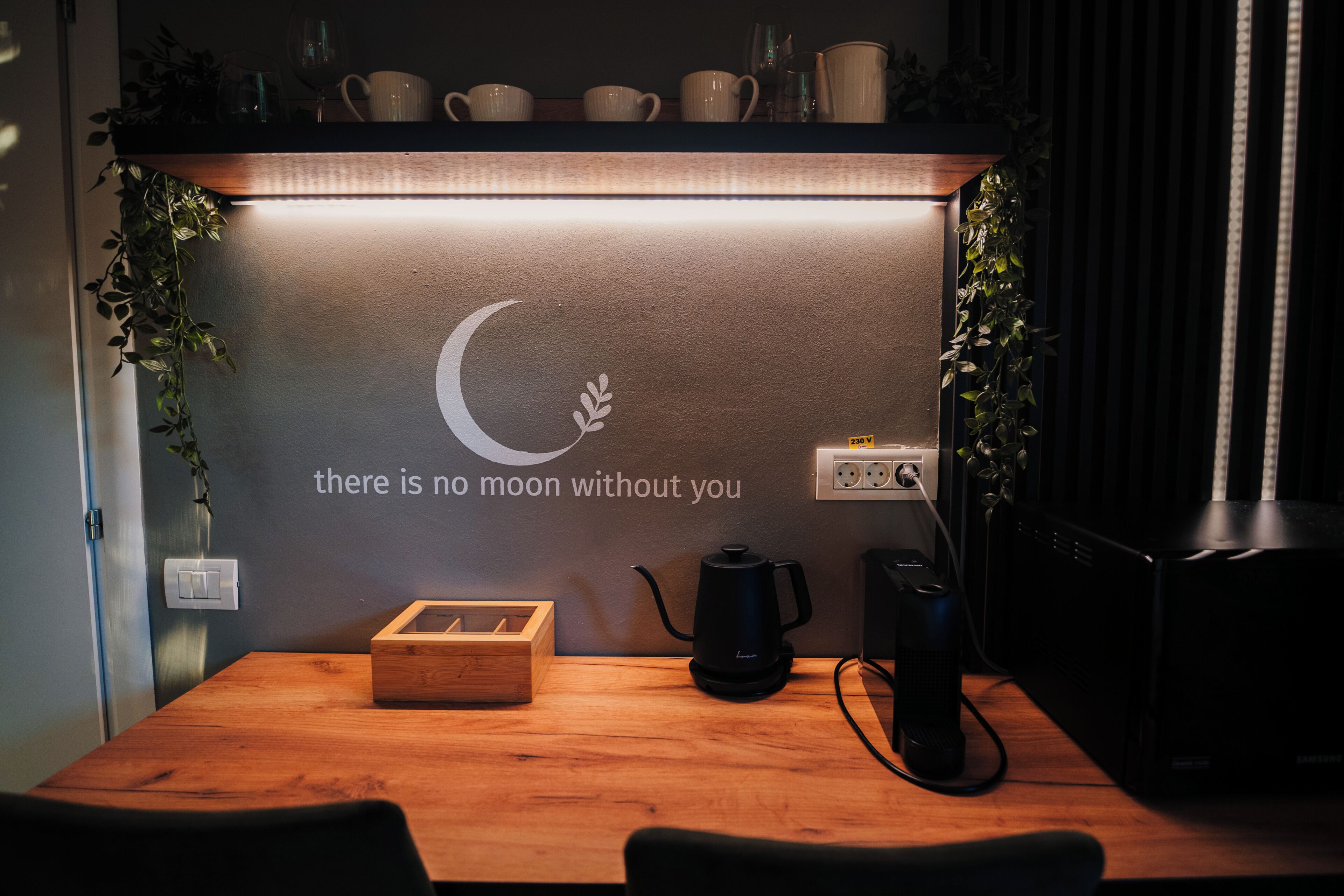 Un espacio de trabajo ordenado con una mesa de madera, una cafetera negra, una caja de madera y una pared con un diseño de luna creciente y una cita motivacional.