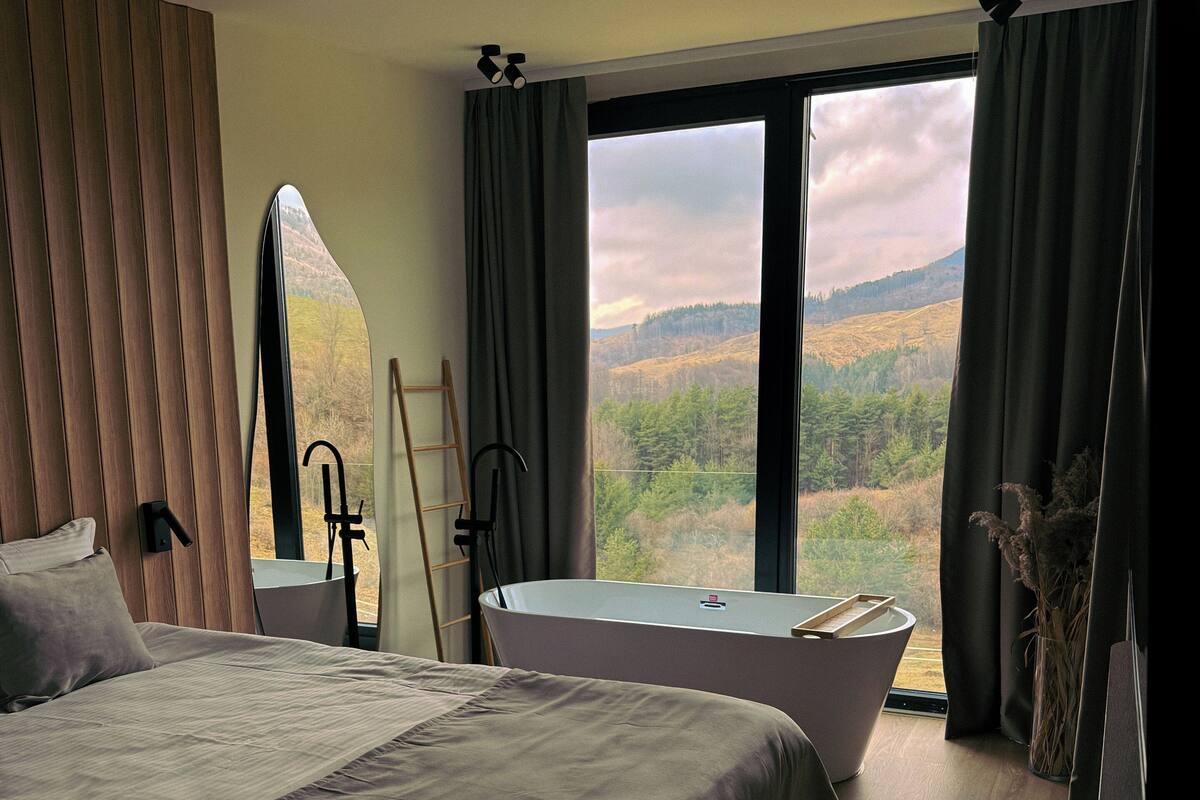 Een slaapkamer met een groot bed, een badkuip, een raam met uitzicht op een bos, en een spiegel.