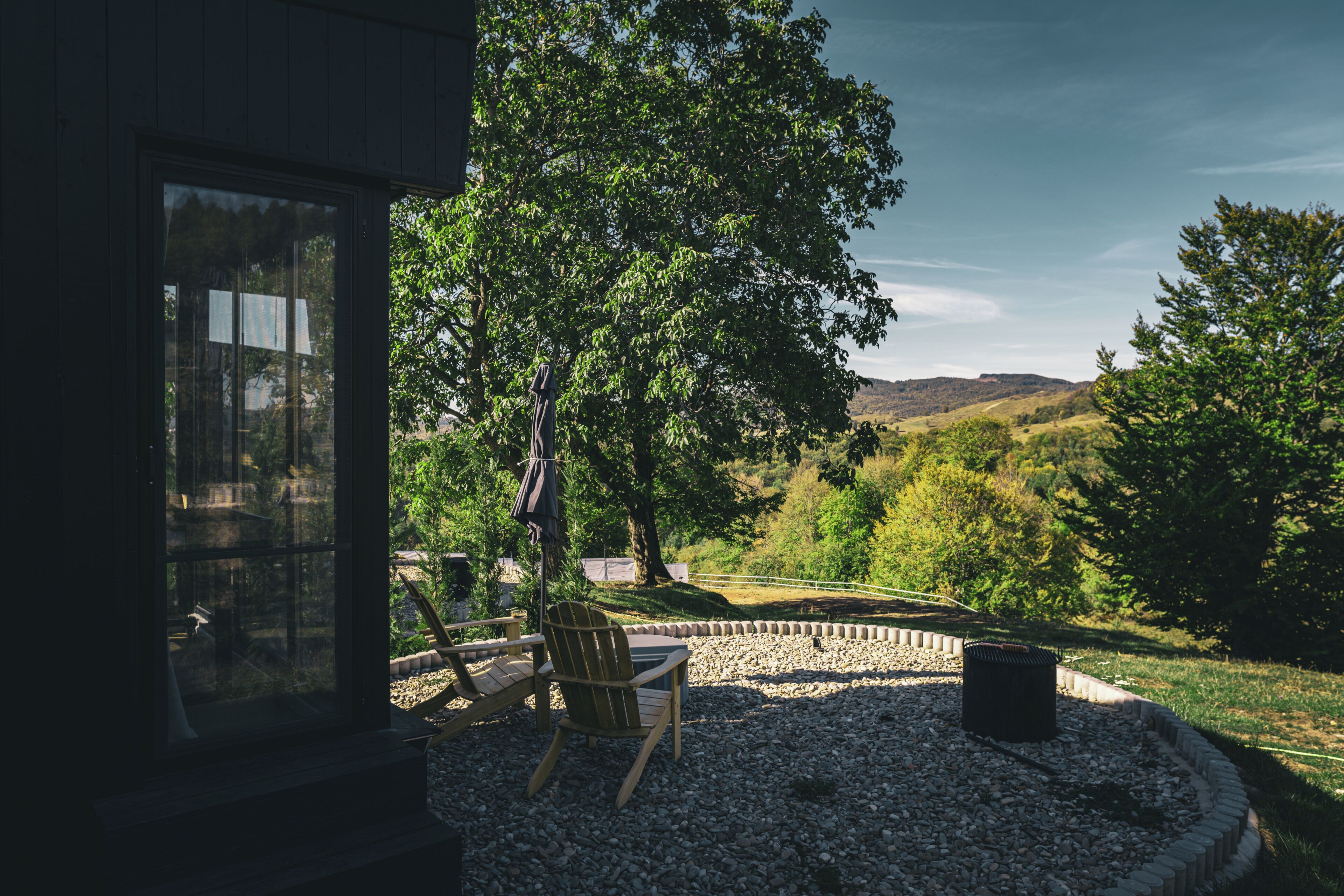 A patio with a table and chairs, a fire pit, and a view of a green landscape with trees and hills.