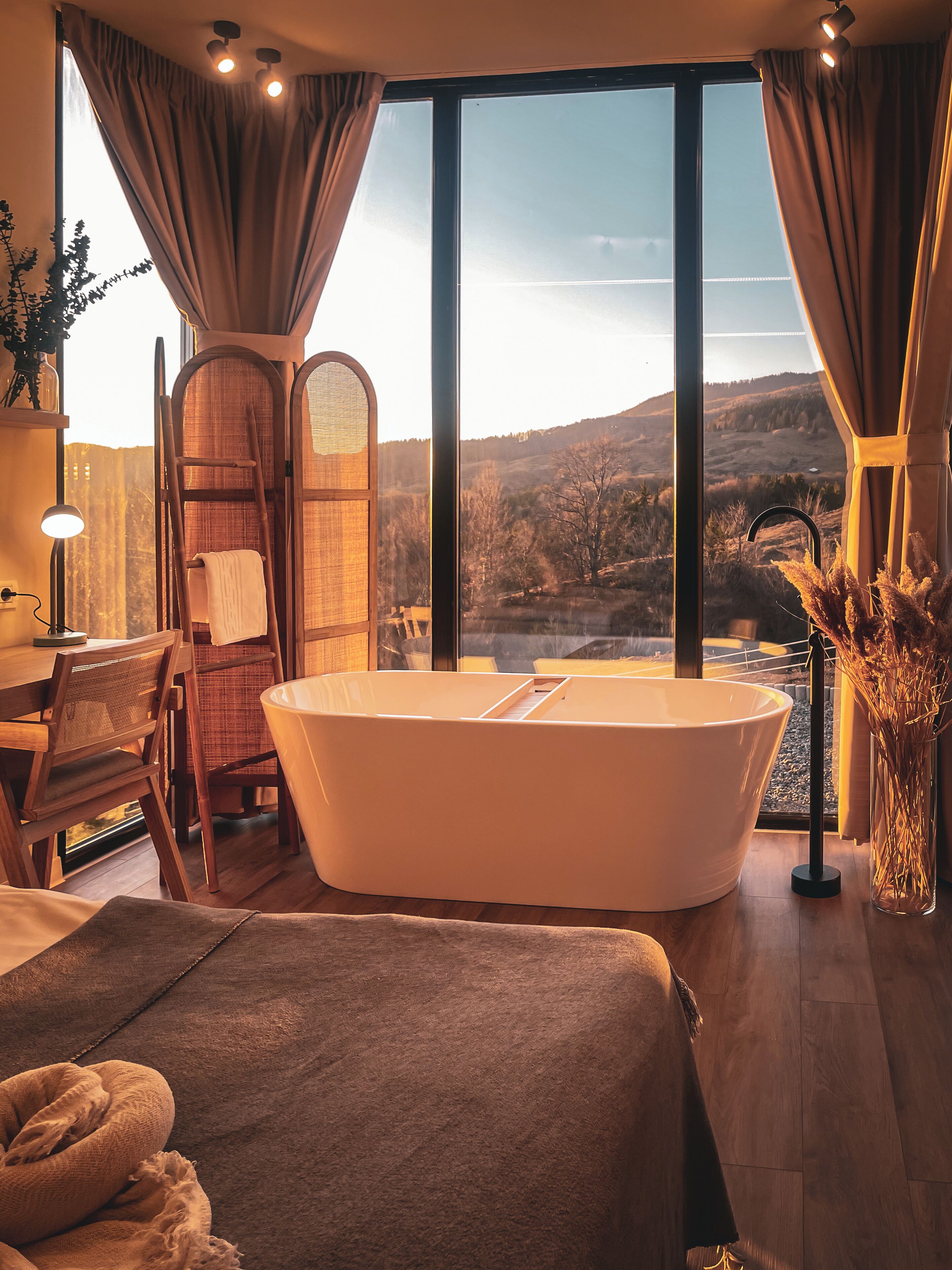 A modern bathroom with a freestanding bathtub, a wooden chair, a large mirror, and a view of a mountainous landscape through large windows.