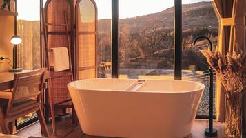 A modern bathroom with a freestanding bathtub, a wooden chair, a large mirror, and a view of a mountainous landscape through large windows.