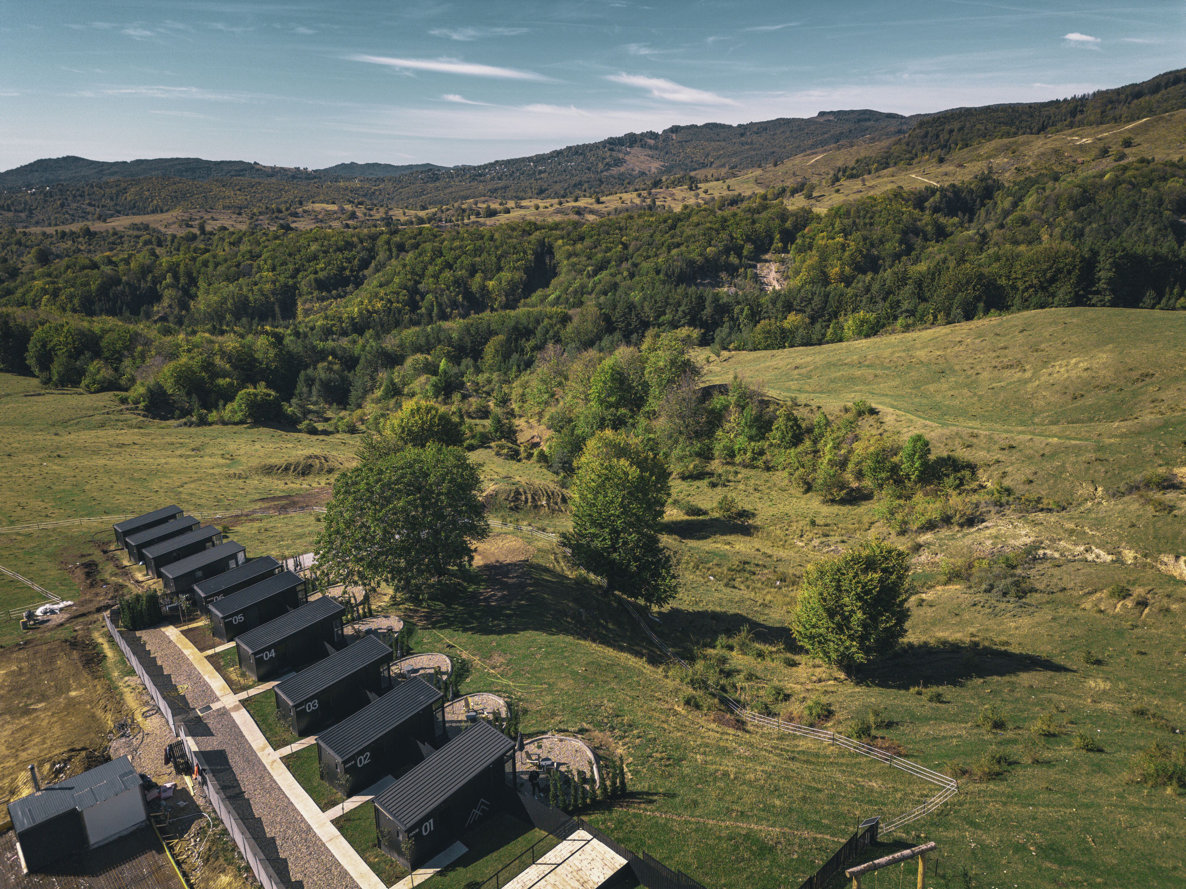 Filera de cabanes modernes de color fosc amb un camí asfaltat i una zona tancada per una tanca, envoltades per un paisatge verd i unes muntanyes al fons.