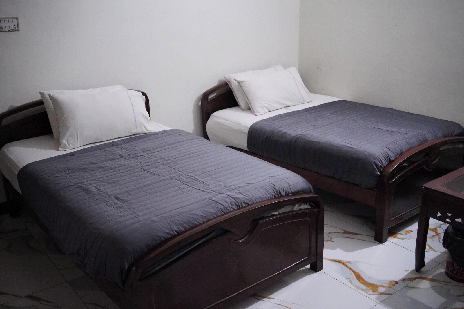 Quarto com duas camas de solteiro, cada uma com estrutura de madeira e roupa de cama branca. O quarto possui piso de azulejo e uma pequena mesa de madeira.