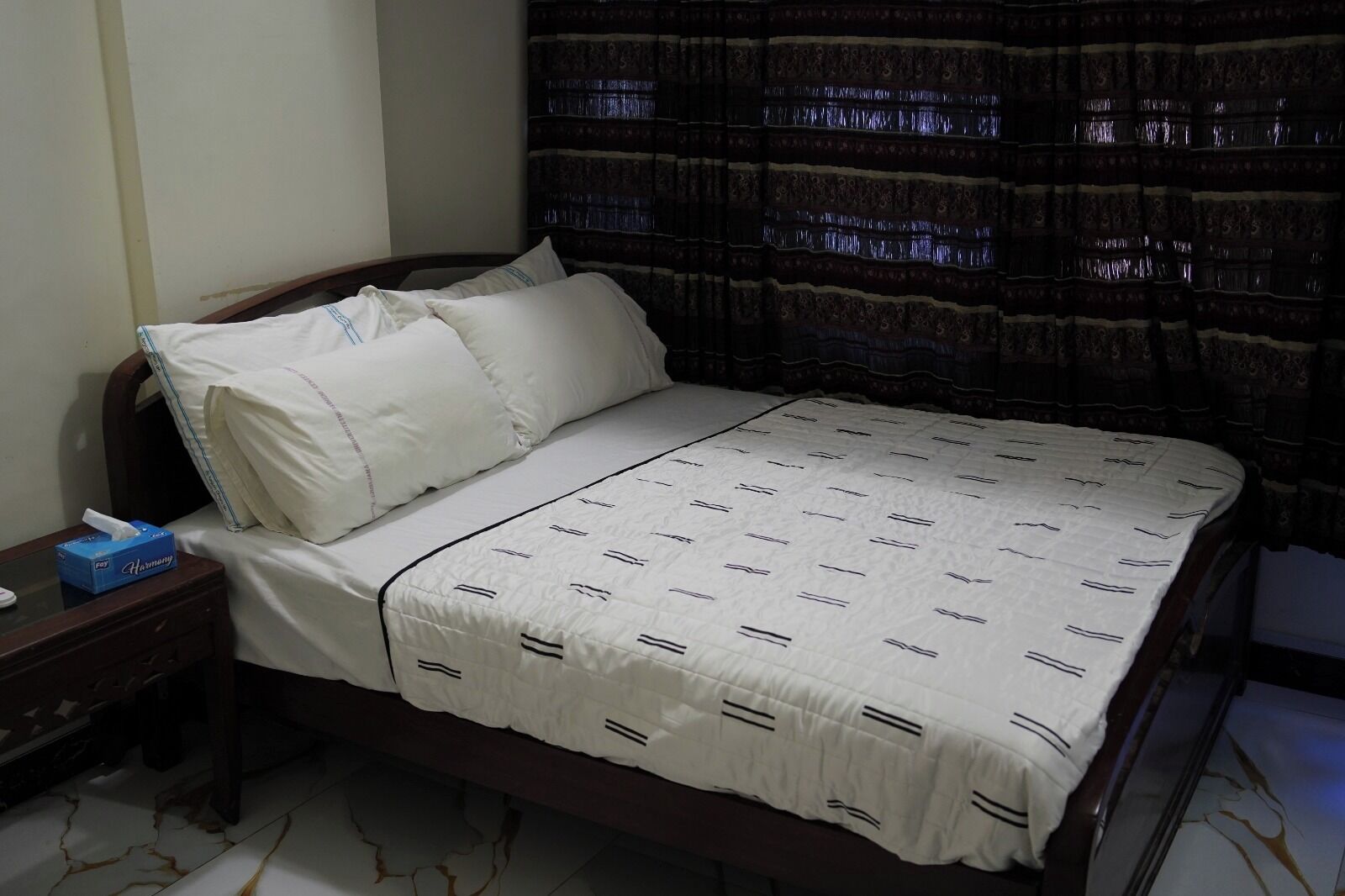 Un dormitorio con cama, almohadas, una mesita de noche y una caja de pañuelos, y una cortina.