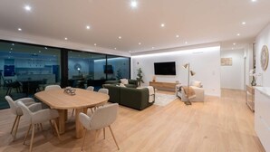 A modern living room with a dining area, a sofa, a TV, and a kitchen visible through glass walls.
