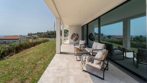 A modern outdoor patio with a sofa, chairs, and a hanging chair, overlooking a scenic hillside.