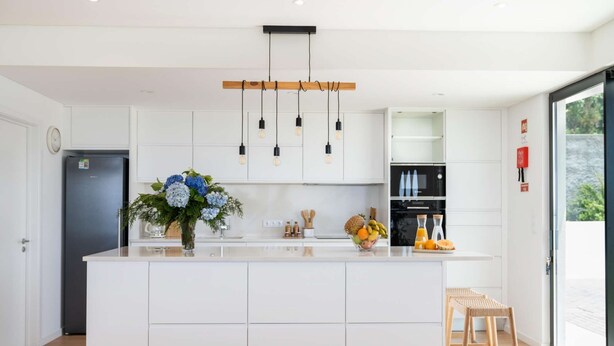 A modern kitchen with white cabinetry, a central island, hanging pendant lights, and a view of the outdoors through a glass door.