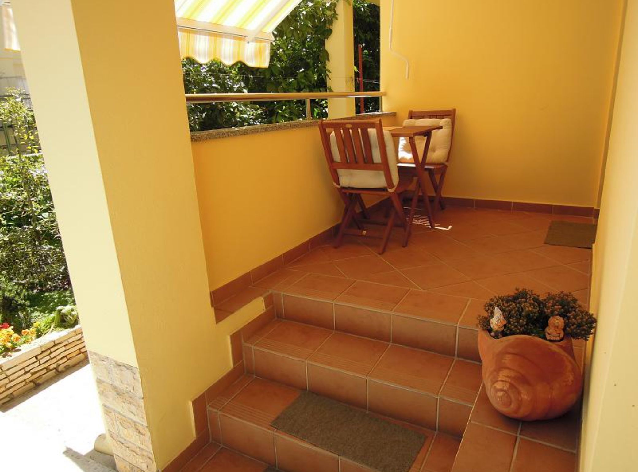 A small balcony with a table and chairs, a potted plant, and a view of greenery.