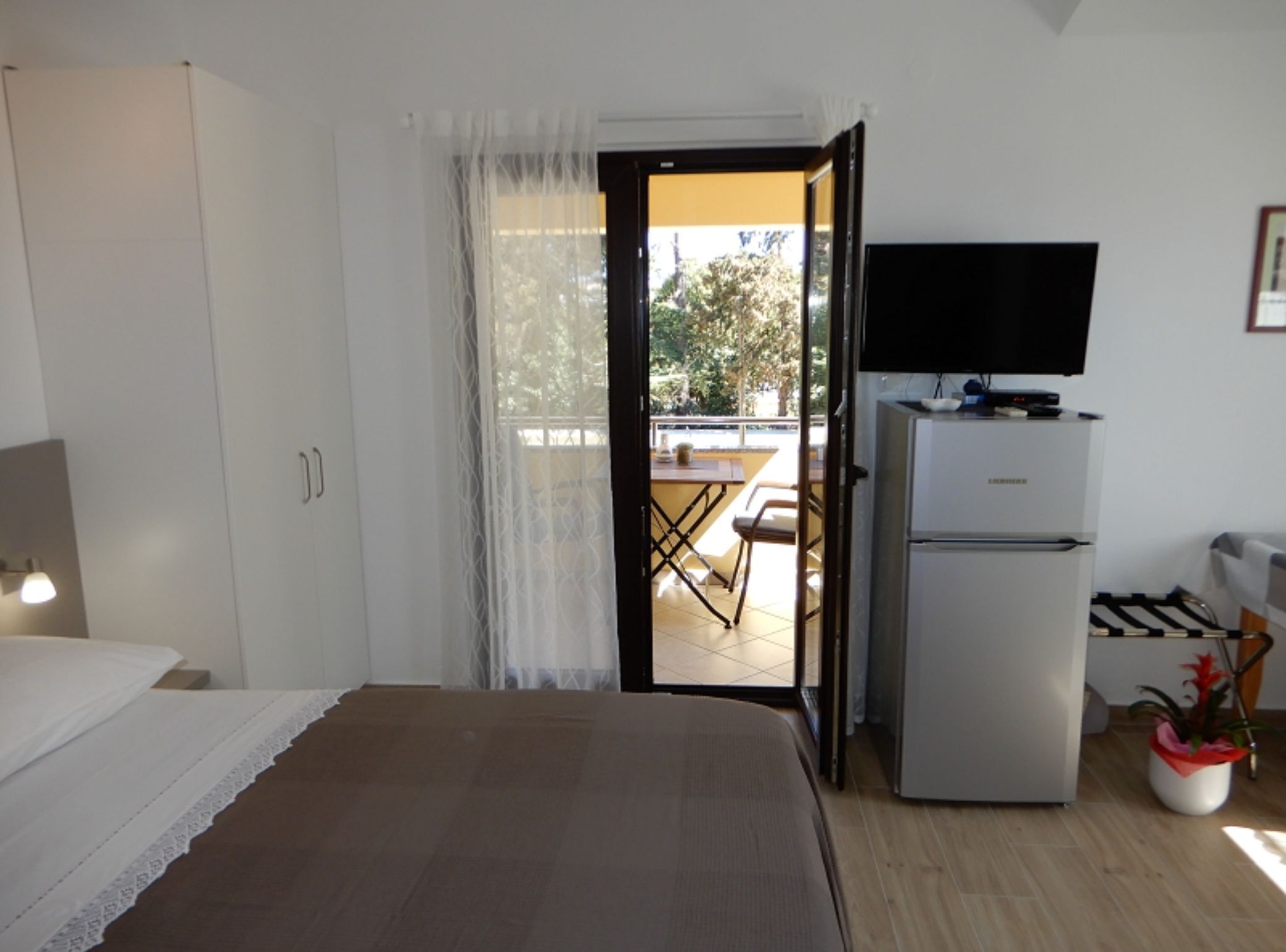 A bedroom with a bed, a television on a stand, a refrigerator, and a balcony with a table and chairs.