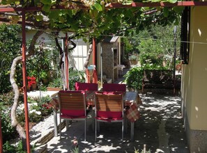 Patio esterno con tavolo e sedie, circondato da vegetazione e un muro in pietra.