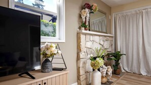 A living room with a flat-screen TV on a wooden cabinet, a stone fireplace, and various potted plants and flowers. - @ Marbella Lane - Nature Escape at Shady Lake (Renton)