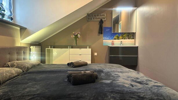Une chambre avec un plafond en pente, un lit recouvert d’une couverture sombre, une commode, un miroir et une petite fenêtre donnant sur une plage.