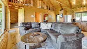 Ein Holzblockhaus-Innenraum mit einem großen, grauen Ledersofa in L-Form, einem runden Holztisch und einer Küchenzeile mit Holzschränken und einem Kühlschrank.