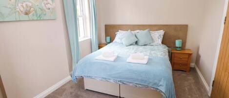 Un dormitorio con cama, mesitas de noche, una ventana con cortinas y un cuadro de flores en la pared.