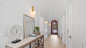 En hall med vita väggar, en träbänk, en stor spegel och en dörr med ett välvt fönster.