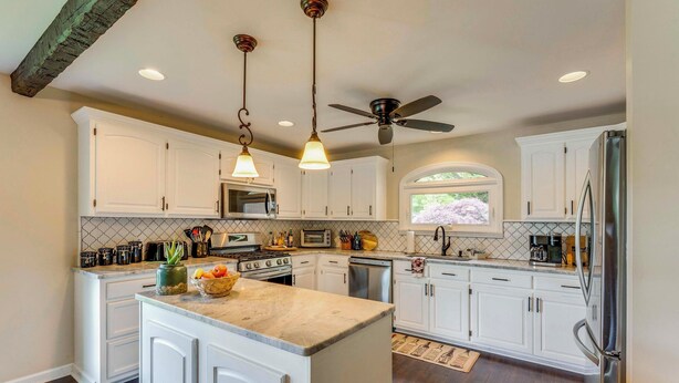 Una cocina con gabinetes blancos, una isla central, electrodomésticos de acero inoxidable, iluminación colgante y un ventilador de techo.