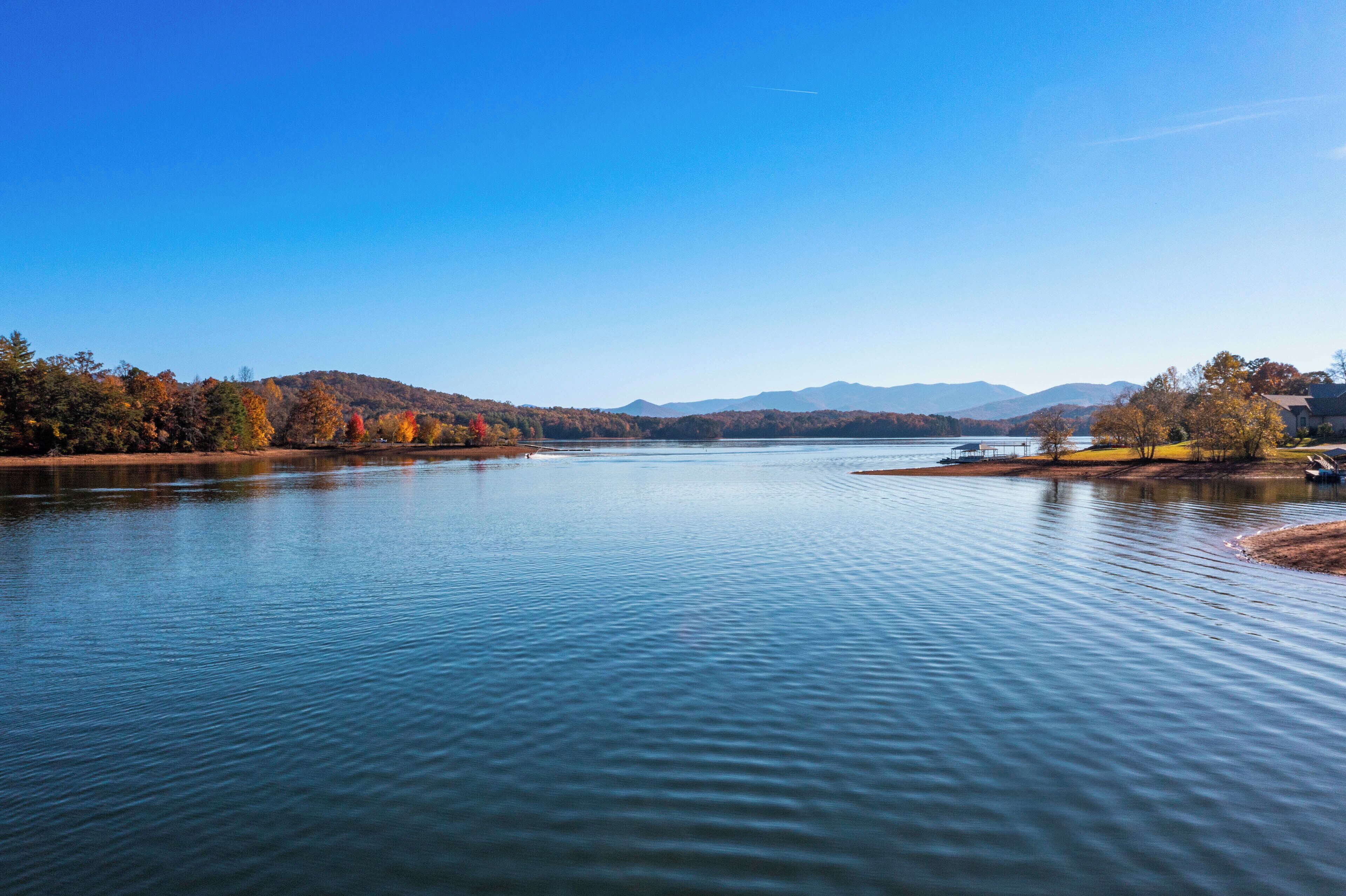 A calm lake with a clear sky, surrounded by autumn-colored trees and distant mountains.