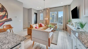 Ein modernes Wohnzimmer mit einer Couch, einem Couchtisch und einem Essbereich. Ein Fernseher ist an der Wand montiert und es gibt ein großes Fenster mit Vorhängen.