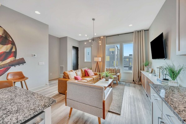 A modern living room with a sofa, coffee table, and dining area. There is a TV mounted on the wall and a large window with curtains. - Rooftop Deck Grill Ping Pong Near Downtown Nashville (Nashville)