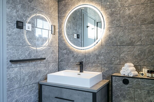 A modern bathroom with a white sink, a round mirror with lighting, a towel rack, and a vanity with toiletries.